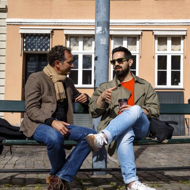 Två män sitter på en bänk. I bakgrunden ett gult hus och en portgång.