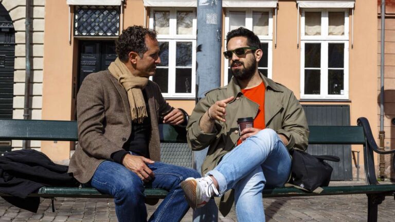 Två män sitter på en bänk. I bakgrunden ett gult hus och en portgång.