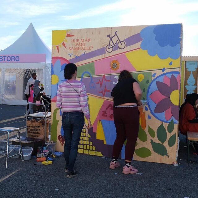 Två unga kvinnor målar ett plank med orden "Hur mår vårt samhälle?"