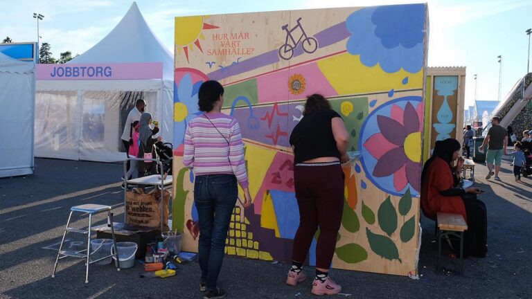 Två unga kvinnor målar ett plank med orden "Hur mår vårt samhälle?"