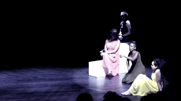 Pitkissä mekoissa pukeutuneita näyttelijöitä tumman näyttämön valokeilassa.