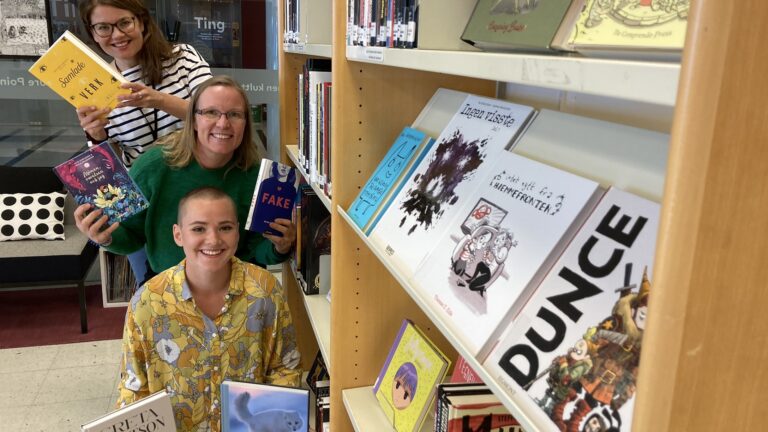 Three smiling librarians holding up books between the bookshelves at the Nordic Culture Point Library