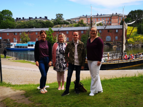 The grants team standing in front of the Suomenlinna harbour. Old boats and a pink building in the background.