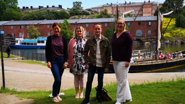 The grants team standing in front of the Suomenlinna harbour. Old boats and a pink building in the background.
