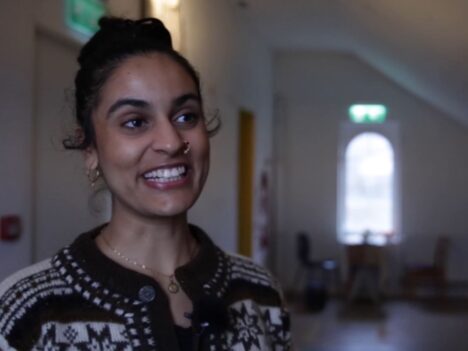 Taiteilija Nayab Ikram hymyilee kameralle, taustalla ikkuna.