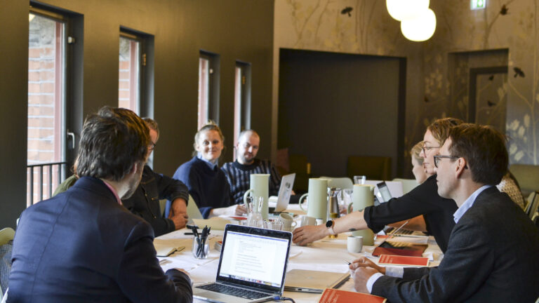Men and women meeting around a table filled with coffee pots and laptop computers.