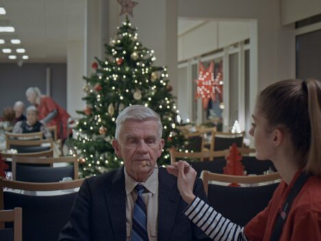 En man blir matad på ett ålderdomshem. Han är iklädd kostym, det står en julgran i bakgrunden.