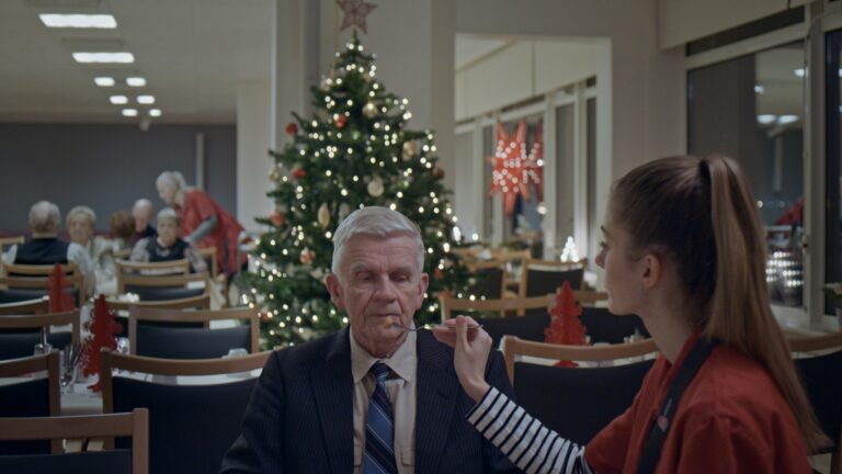 En man blir matad på ett ålderdomshem. Han är iklädd kostym, det står en julgran i bakgrunden.