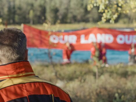 Samisk man tittar mot en protestskylt vid stranden av en älv. Texten lyder "Our land"