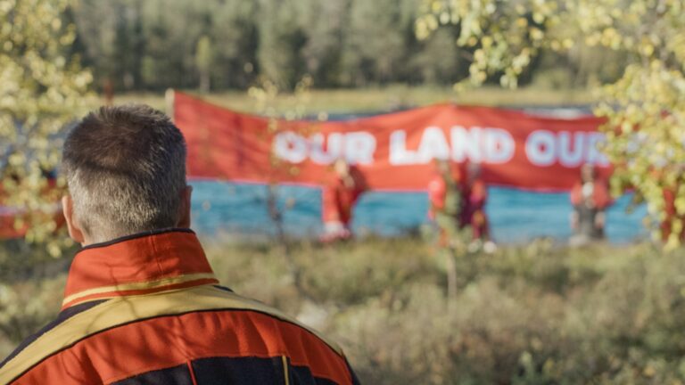 Samisk man tittar mot en protestskylt vid stranden av en älv. Texten lyder "Our land"