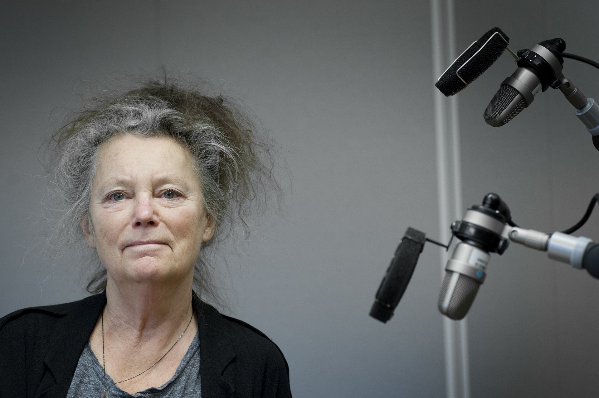 Porträtt av Kerstin Wixe. En äldre kvinna med mörk kavaj och grått hår, hon står bredvid två mikrofoner i en studio