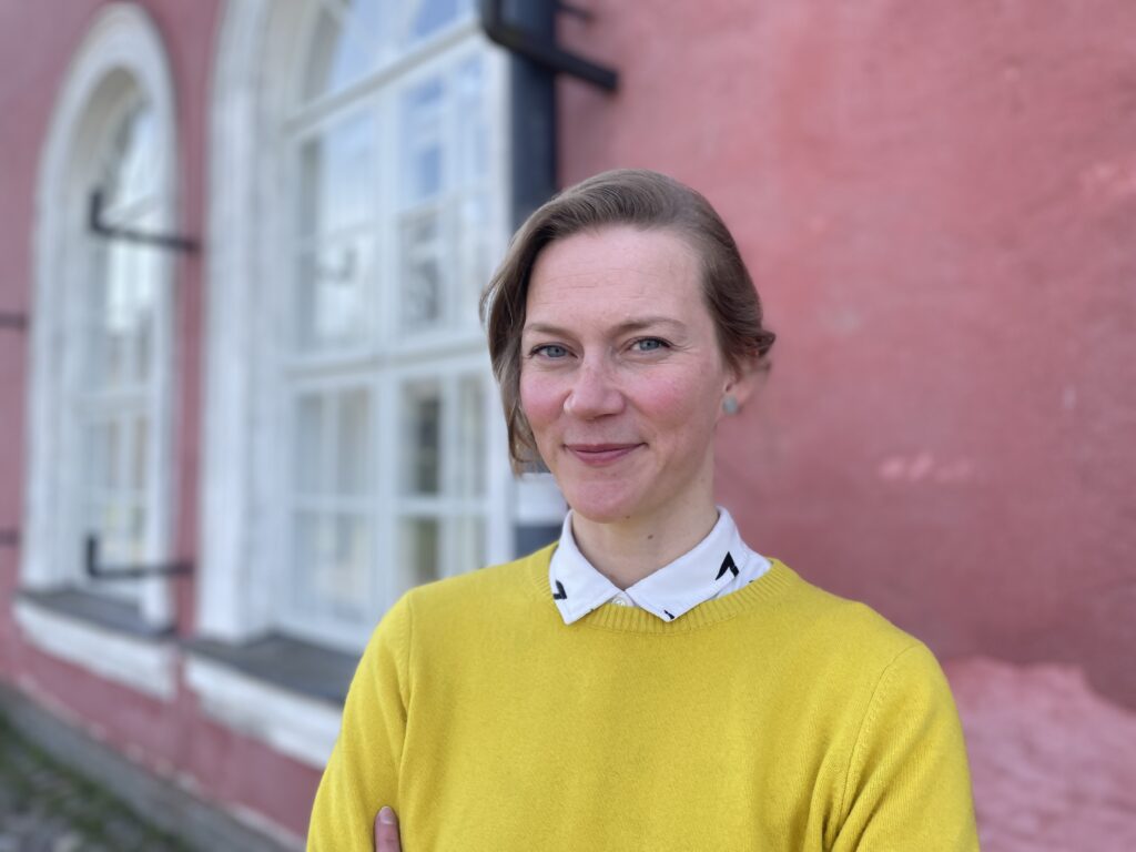 Auri Ahola i gul tröja med en ljusröd husvägg i bakgrunden.