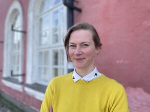 Auri Ahola i gul tröja med en ljusröd husvägg i bakgrunden.