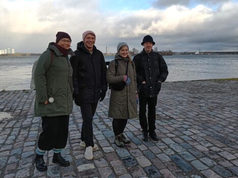 Kaksi miestä ja kaksi naista seisoo rannalla, meri taustalla.