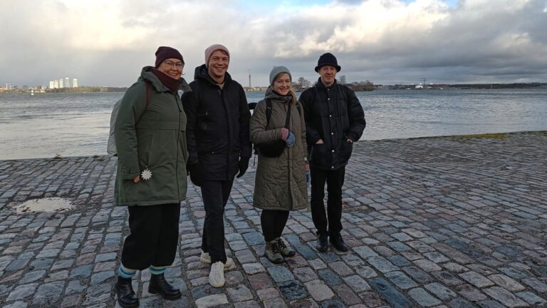 Kaksi miestä ja kaksi naista seisoo rannalla, meri taustalla.