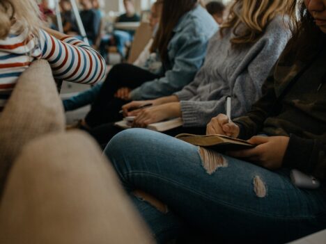 Bild på unga som sitter i en publik. Fokuset är på en ung person med rivna jeans som tar anteckningar i ett häfte.