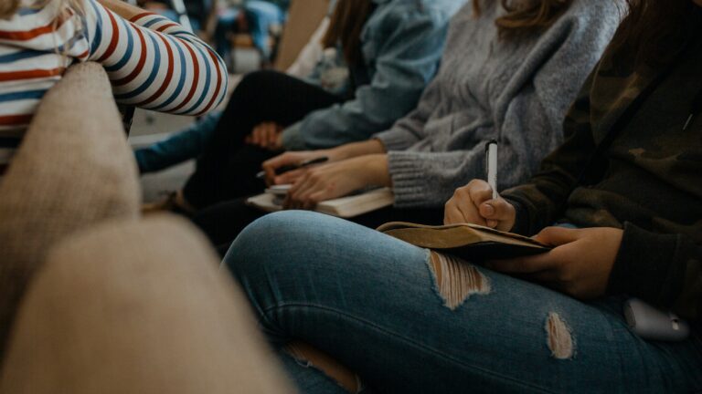 A picture of young people sitting in the audience. The focus is on a young person with ripped jeans taking notes in their notebook.
