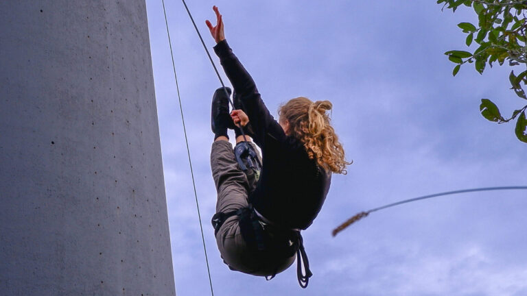 Ett foto av en dans- och cirkusartist med blont lockigt hår som hänger i ett säkerhetsrep, fäst vid en betongbyggnad, och uppträder mot en klarblå himmel. Fotot är taget från marken.