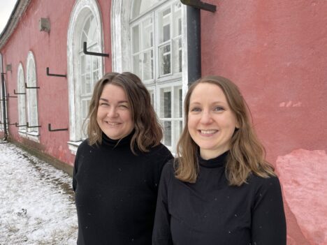 Kaksi naista, joilla on ruskeat hiukset ja musta villapaita seisovat vierekkäin ja katsovat kameraa hymyillen. He seisovat ulkona vaaleanpunaisen kivirakennuksen edessä.