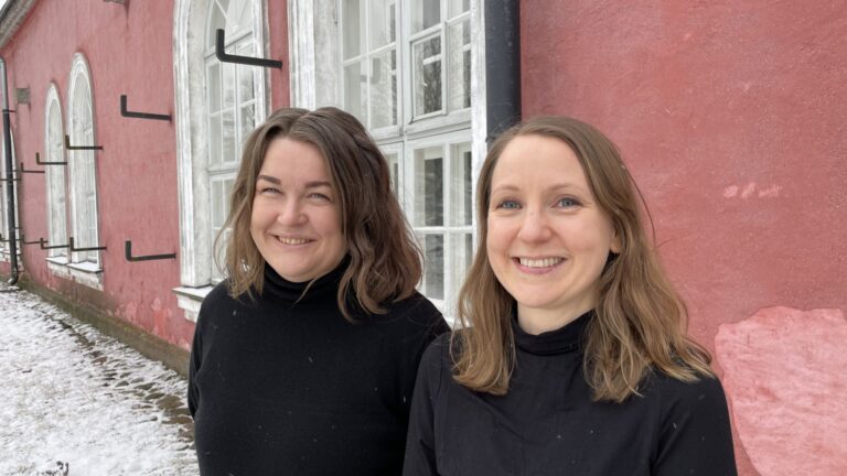 Two women with brown hair and black sweaters stand next to each other, smiling at the camera. They are standing outdoors in front of a light red stone building.