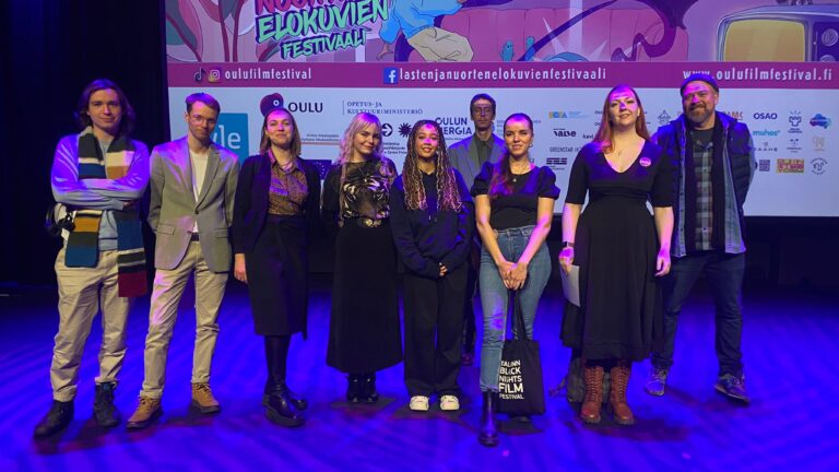 En grupp unga står på en scen och tittar mot kameran. Scenen är upplyst i blått.