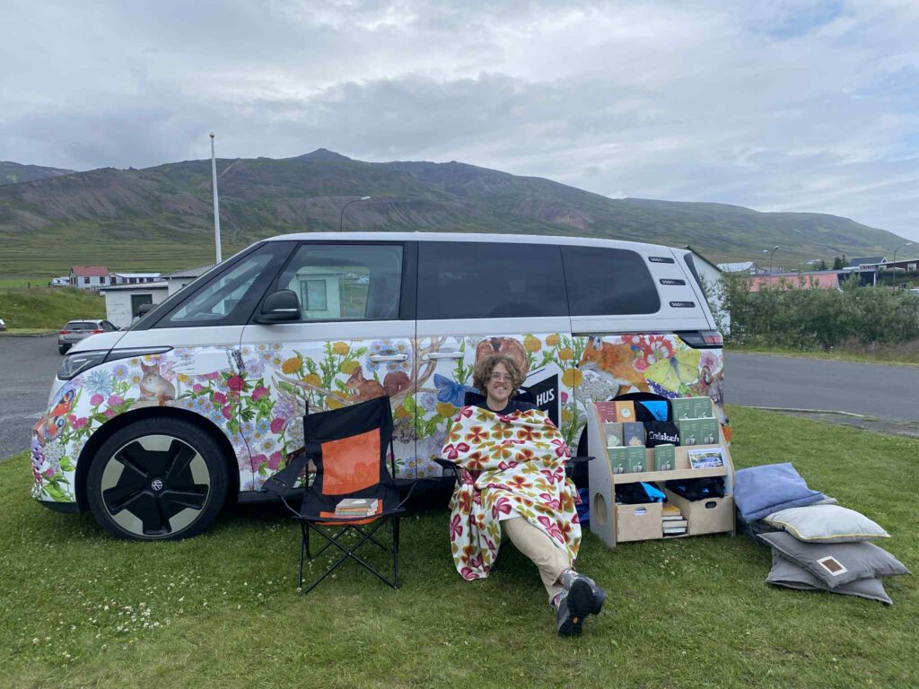 En ung man sitter utomhus i en campingstol framför en bil. Han ler mot kameran och är invirad i en blommig filt. Bilen är vit med dekorativa mönster av blommor och djur. Bredvid den unga mannen står en liten bokhylla med böcker. Landskapet bakom bilen föreställer berg och ängar.