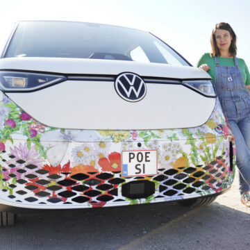 Ruskeatukkainen nainen, jolla on olkapäille ulottuvat hiukset, vihreä paita ja henkselit, seisoo ulkona valkoisen auton vieressä ja katsoo kameraan. Aurinko paistaa, ja auton alaosassa on koristeellisia kukkakuvioita. Auton rekisterikilvessä lukee ”Poesi”.