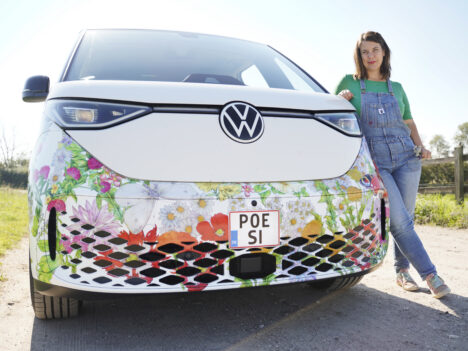 En kvinna med brunt axellångt hår, grön skjorta och hängselbyxor står lutad mot en vit bil utomhus och tittar mot kameran. Solen skiner och den nedre delen av bilen har dekorativa mönster av blommor. På bilens registernummer står det "Poesi".