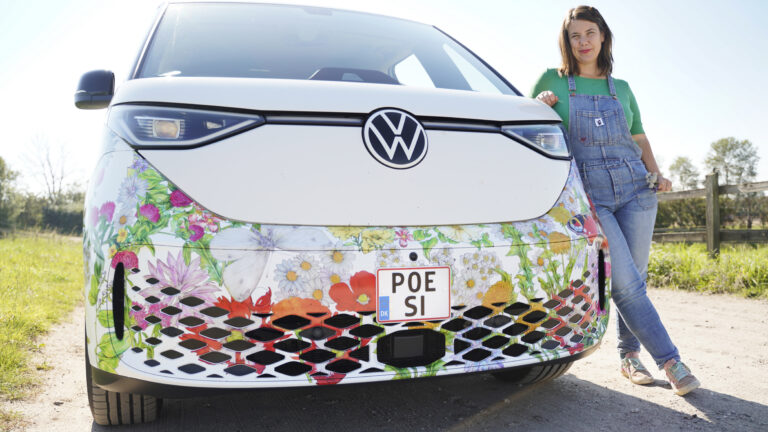 En kvinna med brunt axellångt hår, grön skjorta och hängselbyxor står lutad mot en vit bil utomhus och tittar mot kameran. Solen skiner och den nedre delen av bilen har dekorativa mönster av blommor. På bilens registernummer står det "Poesi".